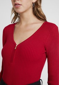 Punainen ribbineulottu bodysuit syvällä V-pääntiellä ja etuvetoketjulla. Pitkät hihat, joustavaa kangasta, jossa hienovarainen kiilto ja teksturoitu kuvio.