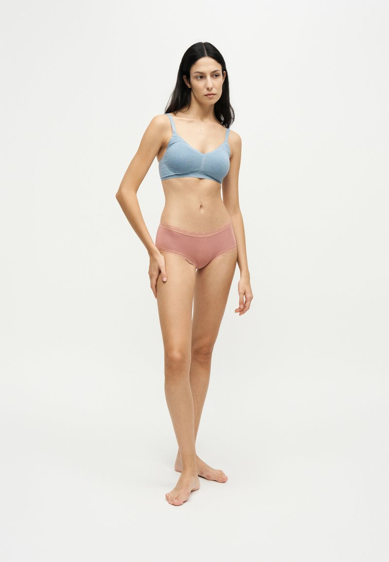 Femme aux longs cheveux foncés, portant un soutien-gorge bleu et une culotte rose bordée de dentelle, debout pieds nus sur un fond blanc uni.