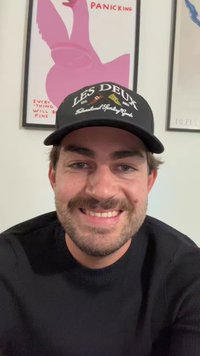 Boné preto com o texto "LES DEUX", usado por um homem com cabelo castanho claro e uma barba, vestindo um suéter preto. Arte de parede colorida ao fundo.