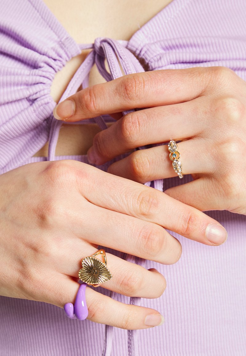 Três anéis nos dedos: um anel de resina lavanda, um anel dourado com um design texturizado de folha e uma banda dourada com várias pedras transparentes.