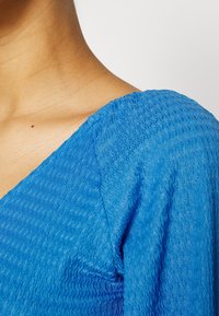 Gros plan sur l'épaule et le cou d'une personne portant un haut en tissu bleu texturé avec un détail froncé à la couture de l'épaule.