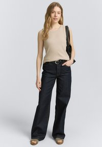Haut sans manches beige, jeans larges en denim noir, ceinture noire et sandales ouvertes à motif. Le mannequin tient un sac à bandoulière noir.