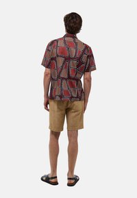 Chemise à manches courtes avec un motif géométrique multicolore dans les rouges et bruns, assortie à un short beige et des sandales noires.