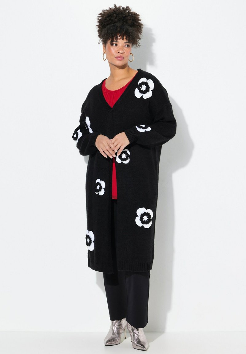 Schwarze Strickjacke mit weißen Blumenmustern, mit V-Ausschnitt und gerippten Bündchen. Über einem roten Oberteil getragen und kombiniert mit glänzenden silbernen Ankle Boots.