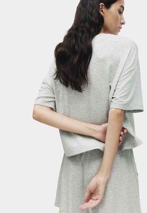 Donna con capelli lunghi e scuri che indossa una maglietta a maniche corte grigio chiaro e pantaloncini abbinati, in piedi con le mani dietro la schiena.