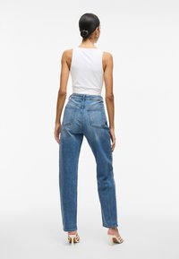Jean en denim bleu taille haute avec une coupe décontractée, présentant des poches arrière et des coutures visibles. Le modèle porte un haut sans manches blanc et des talons.