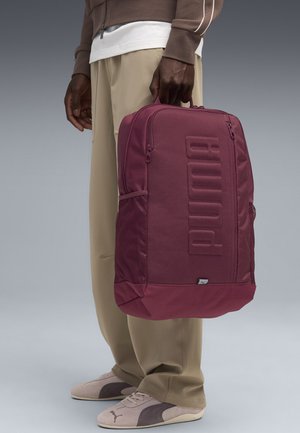 Sac à dos bordeaux présentant un devant texturé avec un logo en relief, une poche latérale en maille et des fermetures éclair. Le matériau est un tissu durable avec une surface lisse.