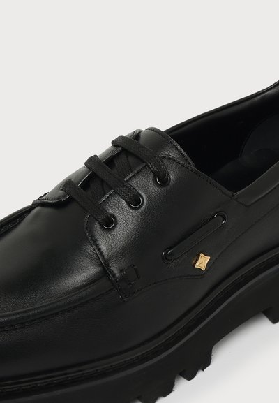 Chaussure en cuir noir à lacets avec semelle épaisse et petit détail carré doré près des lacets sur fond clair.