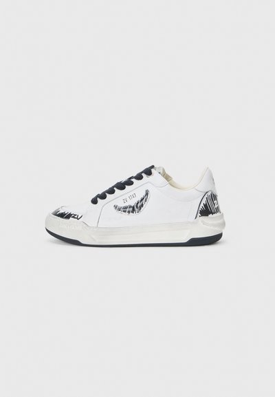 Baskets blanches avec accents noirs, présentant un design texturé sur le côté, des lacets noirs et une semelle en caoutchouc. Logo et illustration sur le dessus.