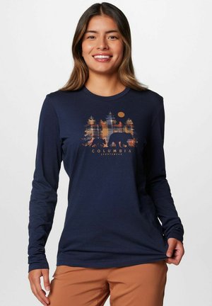 Donna sorridente che indossa una maglietta a maniche lunghe blu navy con orso e cucciolo di orso che camminano tra gli alberi e il logo "Columbia Sportswear", pantaloni beige.