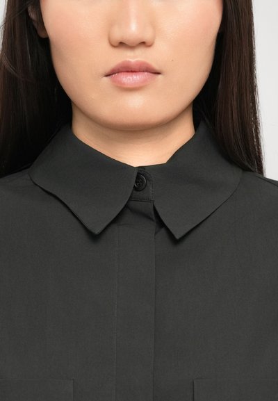 Close-up of a person’s lower face and neck wearing a black collared button-up shirt against a plain background.