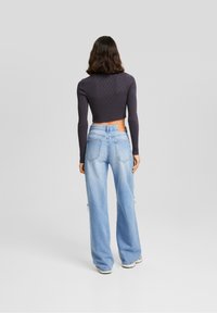 Donna con capelli castani di lunghezza media che indossa un top corto a maniche lunghe scuro e jeans larghi azzurri chiari, in piedi di spalle.