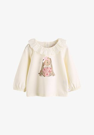 Kremena majica s dolgim rokavi in čipkastim volančastim ovratnikom. Ima ilustracijo zajca z rožami in rožnato obleko. Mehka bombažna tkanina.