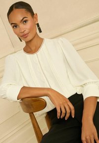 Bluză albă cu mâneci bufante, detalii ondulate și dantelă. Modelul poartă cercei negri dreptunghiulari, așezat pe un scaun de lemn.