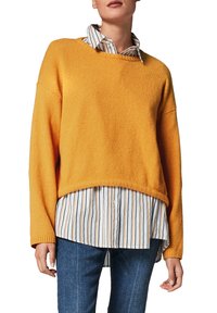 Un pull court jaune vif avec une finition texturée porté par-dessus une chemise à boutons rayée blanche et marine, associé à un jean en denim.