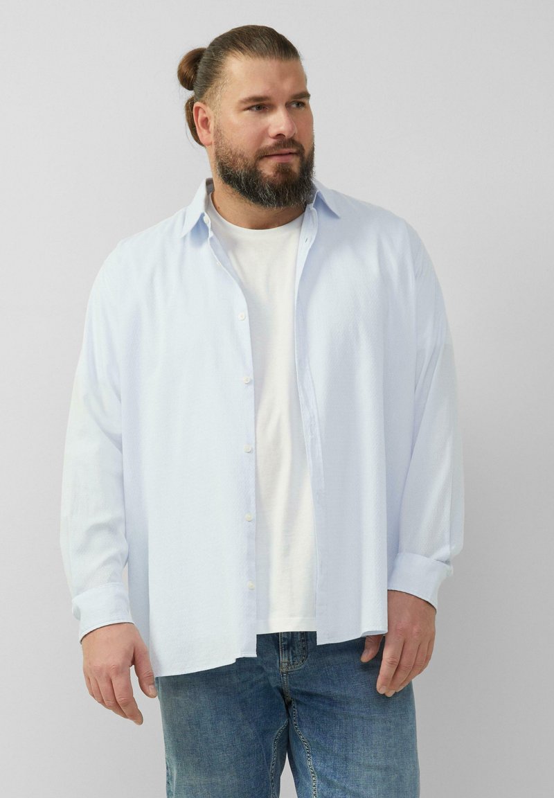 Lichtblauw overhemd met een textuur, open gedragen over een wit t-shirt. Gecombineerd met blauwe spijkerbroeken. Casual ontwerp.