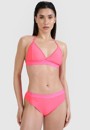 Kobieta z długimi ciemnymi włosami, ubrana w jasnoróżowy halterowy bikini top i dopasowane wysokie majtki bikini na jednolitym tle.
