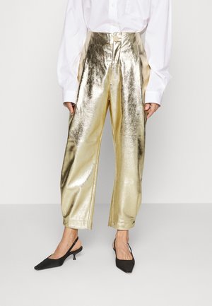 Femme portant un pantalon taille haute métallique doré brillant, une chemise blanche à manches longues, et des talons slingback noirs, debout devant un fond uni.