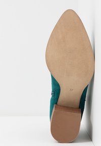 Tummaa turkoosia mokkanahkakenkä, jossa on terävä kärki ja keskikorkea puukanta. Pohja on sileä, mutta siinä on kuvioitu pito, ja siinä on kokomerkintä "37."