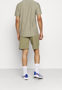 Miestenska športová súprava pozostávajúca z svetlošedej pruhovanej trička a olivovozelených šortiek, doplnená bielymi a modrými teniskami.