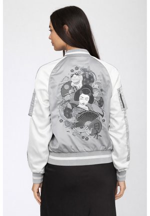 Donna che indossa una giacca bomber grigia e bianca con motivi giapponesi ricamati, tra cui una geisha, pesce koi, ventaglio e gatto portafortuna.