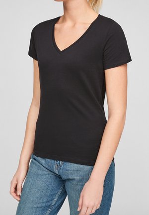 Femme portant un t-shirt noir à manches courtes et col en V, associé à un jean en denim bleu, debout devant un fond neutre.
