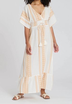 Kvinde iført en hvid maxikjole med beige geometriske mønstre, korte vide ærmer, snøre i taljen og fladflettede sandaler.