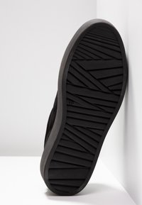 Semelle de chaussure noire avec des motifs géométriques en zigzag et des rainures linéaires, inclinée contre une surface blanche d'angle.