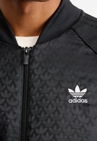 Svart bomberjacka med dragkedja, med ett subtilt ton-i-ton mönster och vit Adidas-logga på vänster bröst. Ribbad halslinning och muddar.