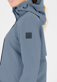 Ljusblå vattentät jacka med huva, med en svart logotyp patch på vänster ärm och minimalistiska design detaljer.