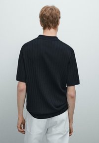 Svart kortärmad polo med ribbad textur och krage. Baksidan har en lätt rundad fåll och en avslappnad passform. Bärs med vita byxor.
