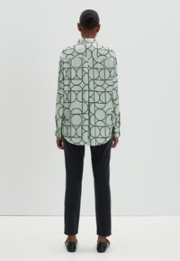 Camicia verde chiaro a maniche lunghe con un motivo circolare nero, caratterizzata da un orlo arrotondato, abbinata a pantaloni scuri e scarpe nere.