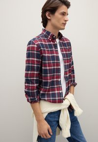 Herren-Flanellhemd in Marineblau, Rot und Weiß kariert, mit Knopfkragen und hochgekrempelten Ärmeln. Weißer Strickpullover um die Taille gebunden.