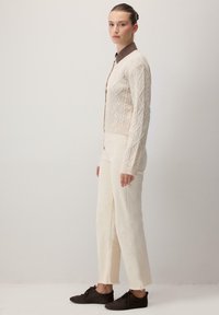 Cardigan crem cu model în diamond, asortat cu pantaloni albi cu talie înaltă și pantofi maro închis. Modelul stă de partea.