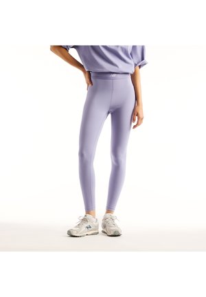 Person trägt hellviolette Leggings mit hohem Bund und ein passendes Oberteil, steht mit einer Hand auf der Hüfte und weißen Sneakers vor weißem Hintergrund.