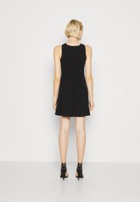 Robe noire sans manches avec un corsage ajusté et une jupe évasée, présentant des plis à la taille. Portée avec des chaussures à talons hauts noires.