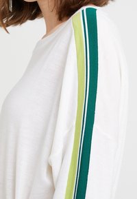 Maglione bianco a maglia con una vestibilità morbida, caratterizzato da strisce verdi e verdi chiaro lungo le spalle, che aggiungono un dettaglio testurizzato al design.