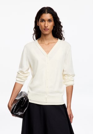 Femme aux cheveux bouclés et foncés portant un cardigan blanc boutonné et une jupe noire, tenant une pochette noire texturée dans sa main gauche.