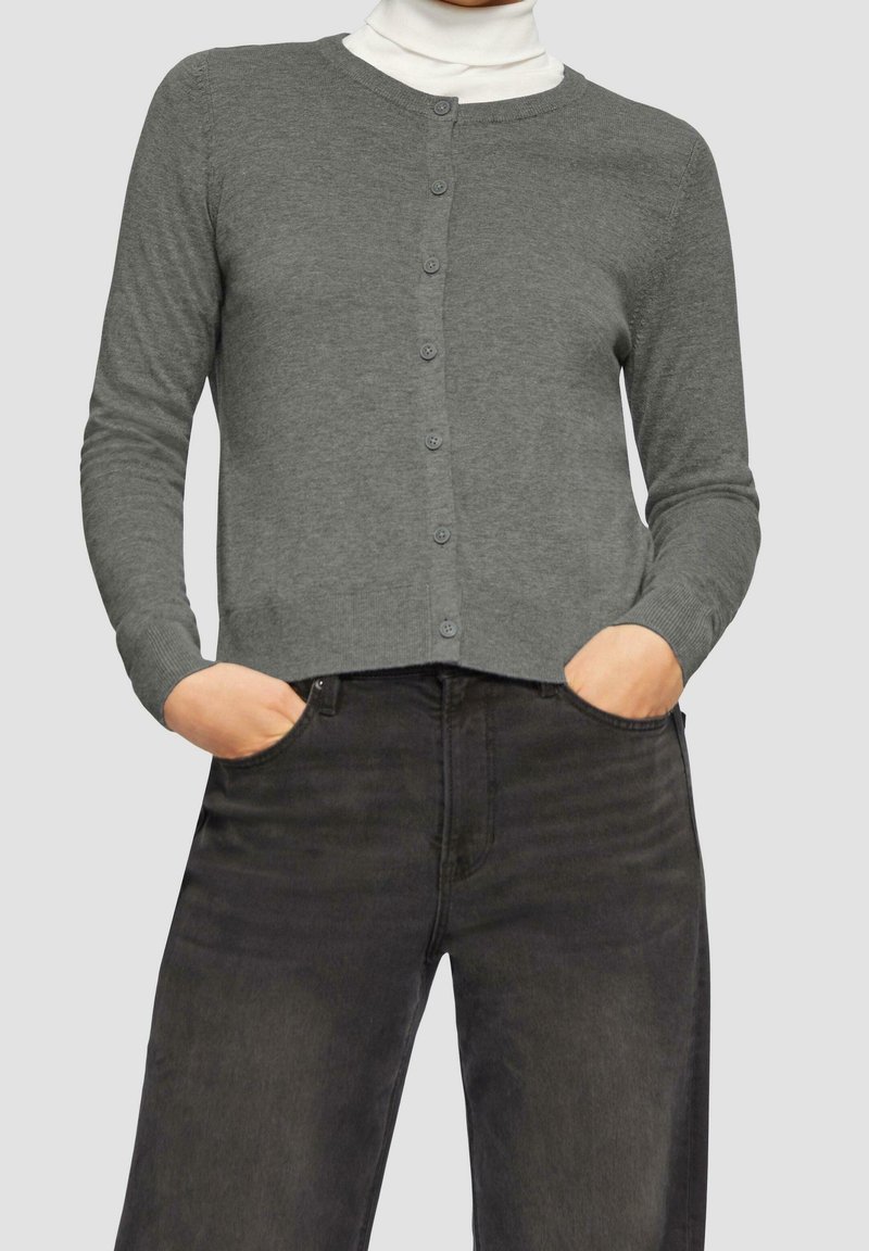 Grauer Cardigan mit rundem Ausschnitt, Knopfverschluss und gerippten Bündchen, kombiniert mit dunklen Jeans. Weicher Stoff und schlanker Schnitt.