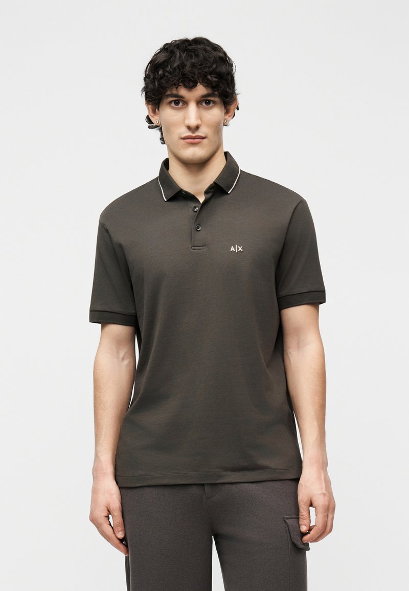 Jeune homme aux cheveux courts et bouclés, portant un polo à manches courtes gris foncé et un pantalon assorti, debout devant un fond uni.