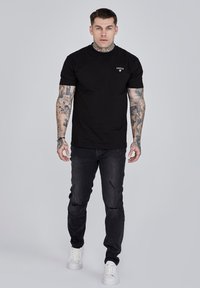 Black short-sleeve t-shirt with a small logo on the chest, paired with distressed black jeans and white sneakers. Flat texture throughout.