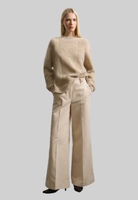 Beige Strickpullover mit einem strukturierten Zickzackmuster, kombiniert mit weitgeschnittenen beigen Hosen aus weichem Stoff.
