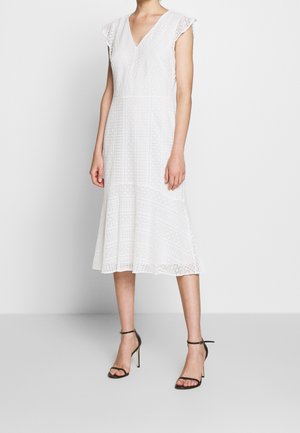 Femme portant une robe midi en dentelle blanche avec des manches courtes griffées et un décolleté en V, associée à des sandales à talons hauts à bride cheville noires.