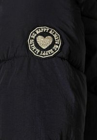 Giacca a vento blu marina con un patch ricamato circolare che presenta un motivo a cuore e il testo "SII SEMPRE FELICE" in oro e nero.