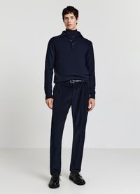 Marineblaues Strickpolo mit hohem Kragen, drei Knöpfen und strukturierter Stoffqualität, kombiniert mit dunklen, plissierten Hosen und schwarzen Schnürschuhen.