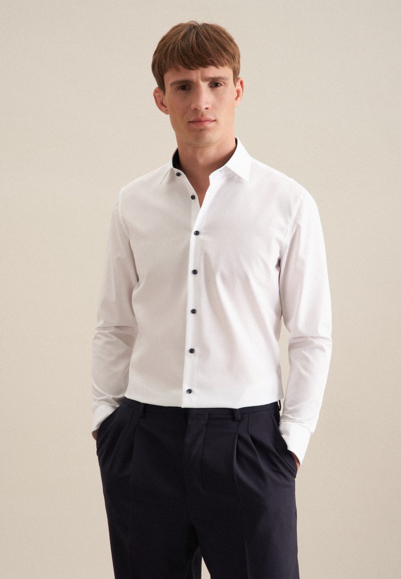 Camicia bianca a maniche lunghe con colletto a punta e bottoni neri, abbinata a pantaloni scuri; il tessuto appare liscio e ben aderente.