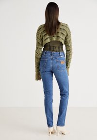 Suéter corto de punto verde oliva con una capa inferior transparente, jeans de tiro alto azules en un lavado claro, que presentan bolsillos traseros y una etiqueta de acento de cuero tostado.