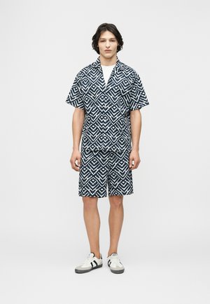 Joven con una camisa de manga corta y pantalones cortos a juego con un patrón geométrico blanco y negro, y zapatillas blancas, de pie contra un fondo liso.