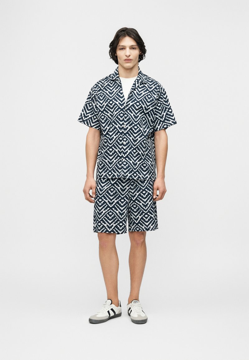 Νεαρός άνδρας φορώντας ένα ταίριαστο σύνολο με μοτίβο γεωμετρικού σχήματος σε μαύρο και άσπρο, με κοντομάνικο πουκάμισο και σορτς, και λευκά αθλητικά παπούτσια, στέκεται μπροστά σε έναν απλό φόντο.