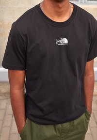 Camiseta negra de algodón con un pequeño logo blanco que dice "The North Face", cuello redondo y mangas cortas. Se lleva con pantalones verdes.